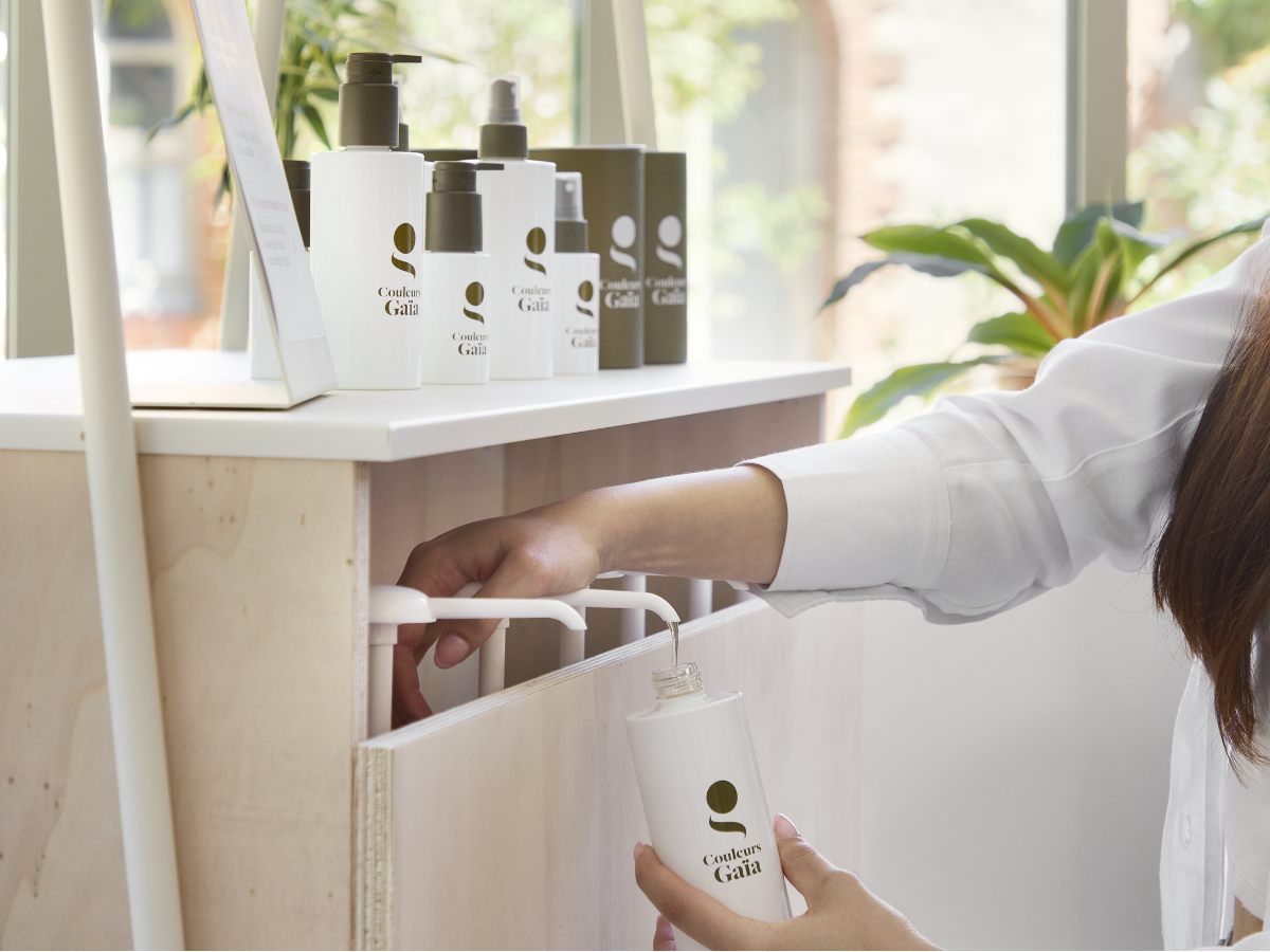 Dans une ambiance de salon de coiffure et pour mettre en avant le vrac chez Couleurs Gaïa, une main tient le récipient rechargeable, l'autre main actionne la pompe du produit pour remplir son récipient.