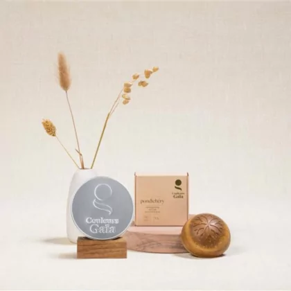 Shampoing Solide Ayurvédique Pondichéry certifié Bio 70g avec son emballage posé sur un socle en bois, ambiance naturelle. Sur un autre socle se trouve la boite adaptée au shampoing 100% aluminium qui a deux fonctions : support dans la douche pour poser le shampoing et transport pour les voyages