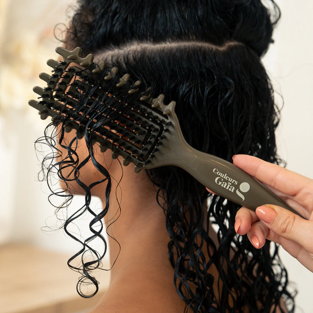 La brosse Couleurs Gaïa pour cheveux bouclés, de couleur kaki avec sérigraphie blanche, est présentée en action en train de définir les boucles de la mannequin