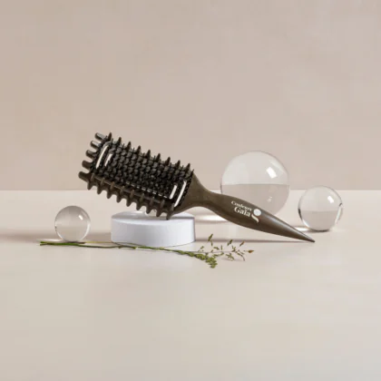 La brosse Couleurs Gaïa pour cheveux bouclés, de couleur kaki, est mise en scène dans un décor naturel et minimaliste. Posée sur un socle blanc, elle est entourée de boules de verre transparentes et de végétaux qui soulignent son design et l'univers naturel de la marque. Ses picots souples et sa forme ergonomique en font une brosse idéale pour démêler les cheveux bouclés sans les agresser.