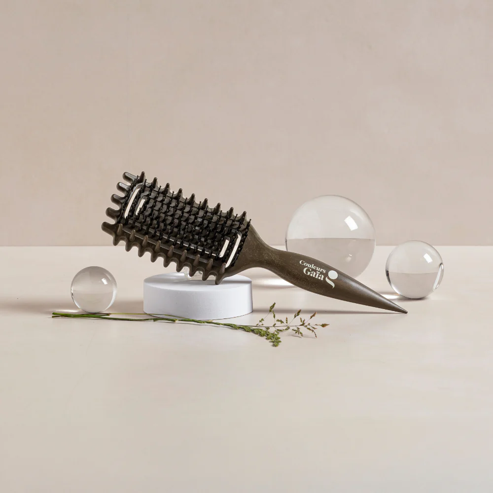 La brosse Couleurs Gaïa pour cheveux bouclés, de couleur kaki, est mise en scène dans un décor naturel et minimaliste. Posée sur un socle blanc, elle est entourée de boules de verre transparentes et de végétaux qui soulignent son design et l'univers naturel de la marque. Ses picots souples et sa forme ergonomique en font une brosse idéale pour démêler les cheveux bouclés sans les agresser.